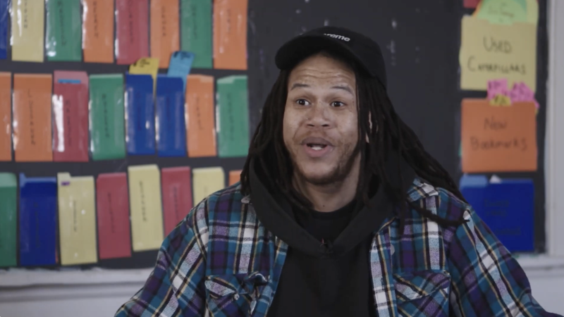 The image shows a man with dreadlocks wearing a black cap, a plaid shirt, and a black hoodie. He is in front of a wall with colorful cards and handwritten notes. The man appears to be talking, and the background suggests an educational or community setting.